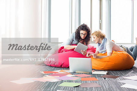 Creative businesswomen using tablet PC while relaxing on beanbag chairs at office