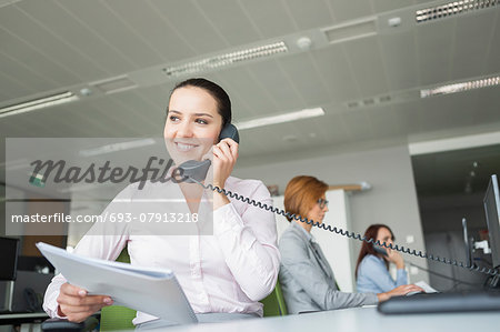 Smiling young businesswoman holding documents while using landline phone with colleagues in background at office