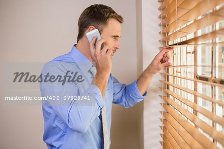 Businessman peeking through blinds while on call in office