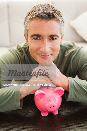Smiling casual man resting head on piggy bank at home in the living room