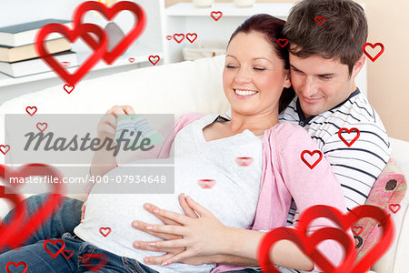 Portrait of a happy pregnant woman holding baby shoes and of her husband on a sofa against hearts