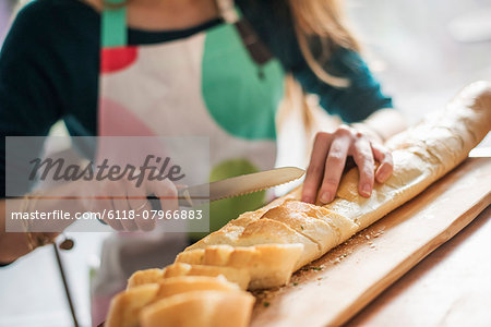 Close up of a woman wearing an apron, sitting at a table, slicing a baguette.