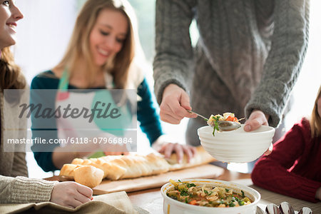 People sitting and standing at a table, a man serving food into a bowl, a woman slicing a baguette.