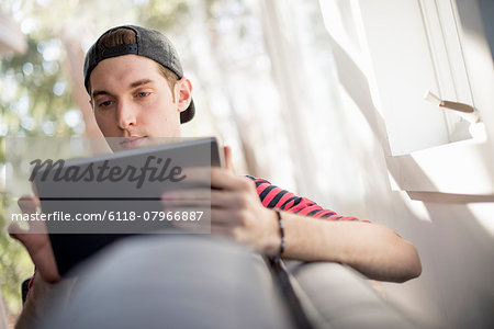 Man wearing a baseball cap backwards, sitting on a sofa, looking at a digital tablet.