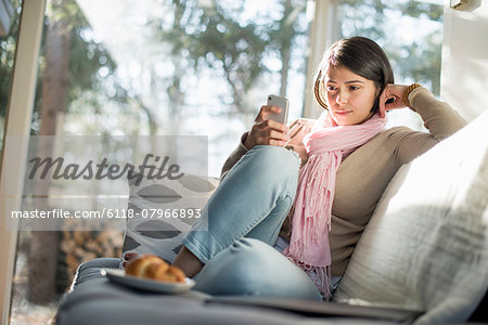 Woman sitting on a sofa looking at her cell phone, a plate with a croissant in front of her.