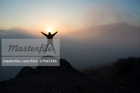 The woman jumps and  lifts her arms in victory. She contemplates beautiful sunset in mountains.