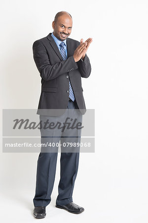 Full length happy Indian businessman in formal suit clapping hands and looking at camera, on plain background.