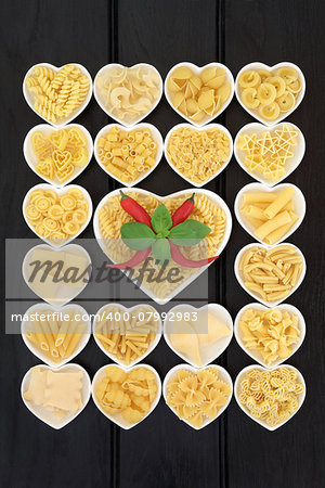 Pasta food selection in heart shaped porcelain dishes with basil herb and red chilli pepper over dark wood background.
