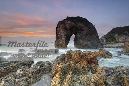 The magnificent natural rock structure, Horse Head Rock, which lies in the beautiful coastal fishing village of Bermagui.  The rock has weathered by the forces of nature over time and resembles a horse drinking water.  Behind the sky lights up in hues of red orange and blue and in the foreground sharp volcanic rocks and rockpools wet with the surging seas