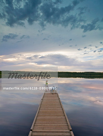 A couple, man and woman sitting at the end of a long wooden dock reaching out into a calm lake, at sunset.