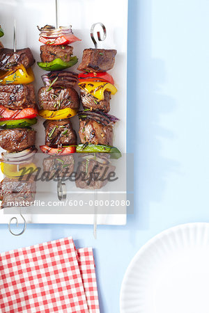 Grilled Beef and Vegetable Skewers on platter with red and white checkered napkins, studio shot on blue background