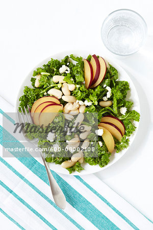 Kale, sliced plum, white bean and feta salad with a blue striped napkin, fork and a glass of water, studio shot on white background
