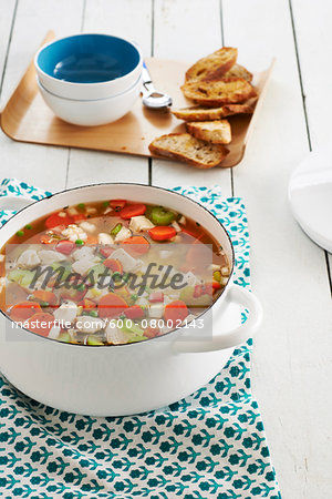 Chicken and vegetable soup in a stock pot with bread and bowls in the background, studio shot