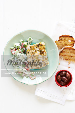 Lemon Cod with Greek Salad on a green plate, with Bread and Olives in bowl on cloth napkin, studio shot on white background