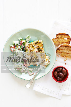 Lemon Cod with Greek Salad and fork on a green plate, with Bread and Olives in bowl on cloth napkin, studio shot on white background