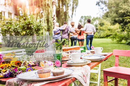 Group of friends walking away with arms around each other, garden party table with food in foreground