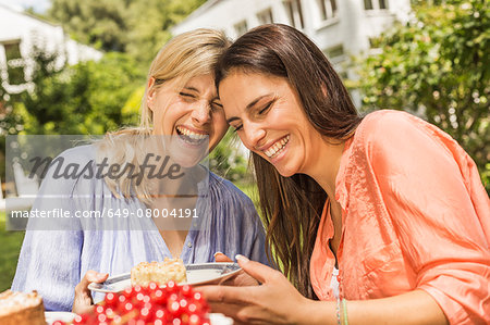 Two female friends in garden, laughing, holding plate with dessert