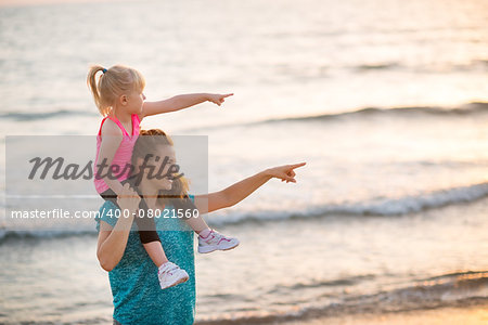 Happy baby girl sitting on shoulders of mother on beach in the evening and pointing