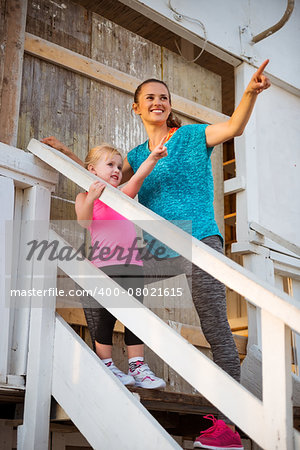 Healthy mother and baby girl standing on stairs of beach house and pointing