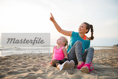 Healthy mother and baby girl sitting on beach in the evening and pointing