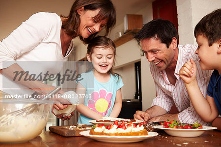 A family, two parents and two children in the kitchen icing a cake with fruit and cream.
