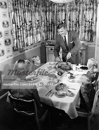 1950s FAMILY OF 5 THANKSGIVING HOLIDAY MEAL MOM DAD MAN WOMAN 3 KIDS BOYS GIRLS AT DINING ROOM TABLE FATHER CARVING TURKEY