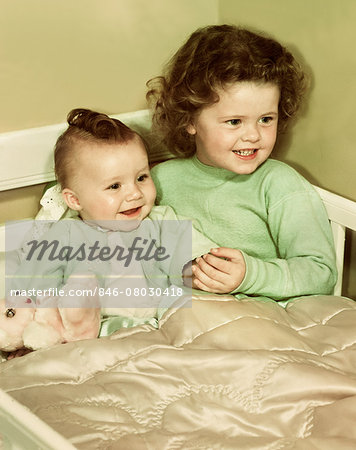 1940s 1950s SMILING LITTLE GIRL AND HAPPY BABY SISTER SITTING TOGETHER IN CORNER OF CRIB BED