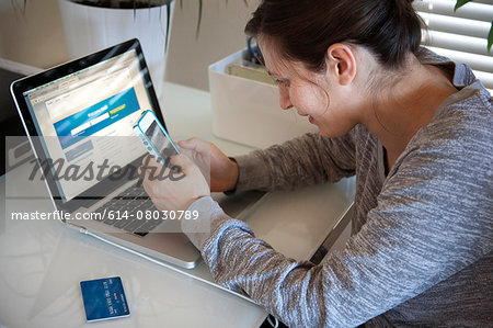 Young woman sitting at table using smart phone and laptop, credit card on table