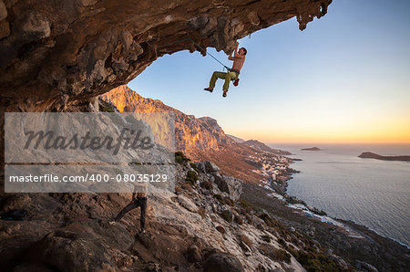 Male rock climber climbing on a roof in a cave, his partner belaying