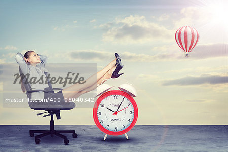 Businesswoman sitting in office chair with her hands clasped behind her head and her feet up on alarm-clock. Sky with clouds and air balloon as backdrop