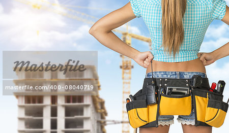 Woman in tool belt with different tools standing backwards, akimbo. Cropped image. Building under construction in background