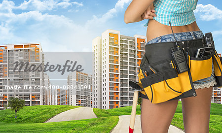 Woman in tool belt with different tools stands back. Cropped image. Green hills with road and buildings on background