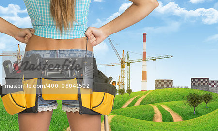 Woman in tool belt with different tools stands back. Hands on hip. Cropped image. Green hills and cranes with heat power stations on background