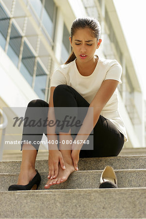 Portrait of latina woman walking on high heels and feeling pain, massaging feet with hand and sitting on stairs