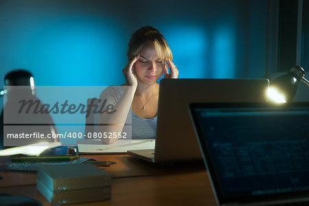 Beautiful woman working as interior designer, staying late at night in office with drawings and laptop computer. The girl feels tired and her eyes hurt.