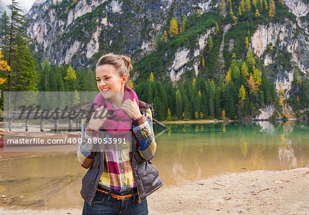 A fit, sporty brunette smiling and readjusting her scarf on the shore of Lake Bries. The great outdoors. The lake reflects the trees and mountains. In the background, a wooden boat and pier.