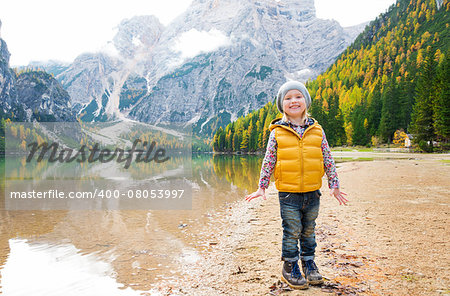 A blonde girl wearing hiking gear stands smiling on the shore of Lake Bries. In fall colours, the mountains, trees, and sand are all reflected in the water.