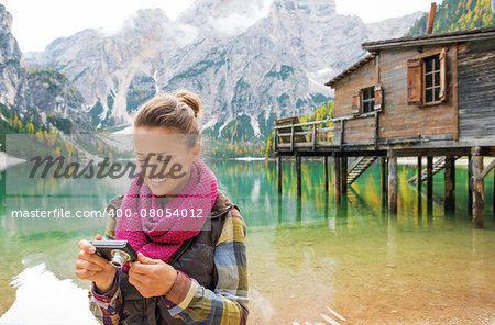 A smiling brunette wearing outdoor gear looks at the photo she has taken on her digital camera. In the background, the Italian Dolomites are above the lake. The still water provides a mirror reflection of the fall colours, wooden pier, and rustic wooden house on the pier.