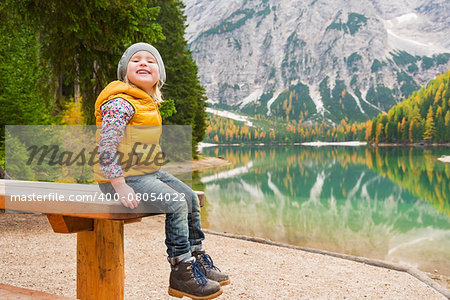 A smiling blonde girl tilting her head back is sitting on a wooden picnic table at Lake Bries, resting. She is wearing outdoor gear. In the background lake water, the mirror and autumn colours are all reflecting in the still water.