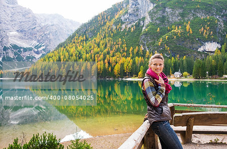 A smiling brunette hiker at Lake Bries wearing outdoor gear is resting against a wooden railing, holding her pink scarf. The still water, autumn colours and shades of green and gray create an autumn background. The water provides a mirror image of the scenery.