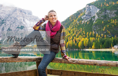 A smiling brunette sits on a wooden guardrail. In the background, autumn colours and the Dolomite mountains are reflecting in the still water of Lake Bries. Golden fall colours.
