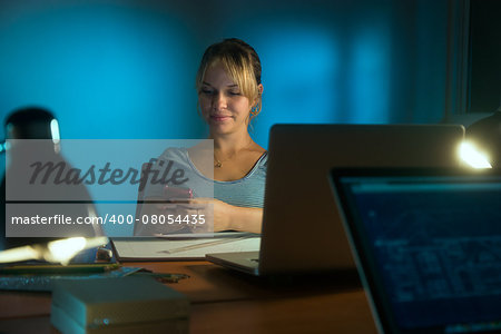 Beautiful woman working as interior designer, staying late at night in office with drawings and laptop computer to complete a project. The girl types a message on mobile phone.