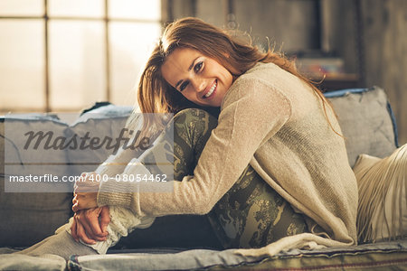 An elegant brunette woman is smiling, hugging her knees. Wearing comfortable, casual clothing, leggings, and a cardigan, she is relaxing on a loft sofa. Industrial chic ambiance and cozy atmosphere.