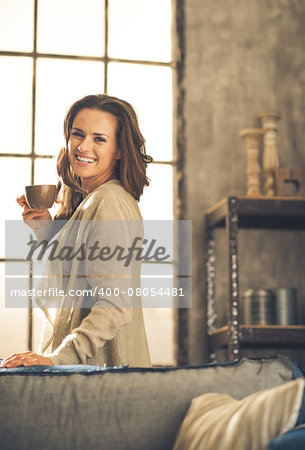 A brunette woman in comfortable clothing is smiling over her shoulder, holding up a hot cup of coffee. Industrial chic background, and cozy atmosphere. Loft decoration details. Upper body shot.