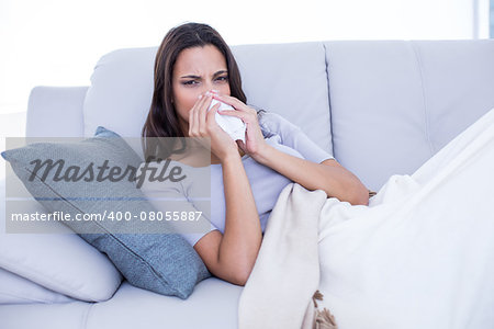 Sick brunette lying on the couch and blowing her nose in the living room