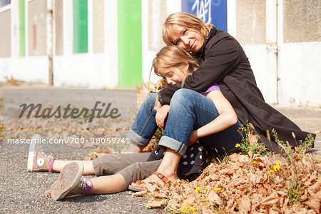 Portrait of mother and daughter sitting on curb on sidewalk in autumn, painted doors of abandoned stables in background, Vendome, Loir-et-Cher, France
