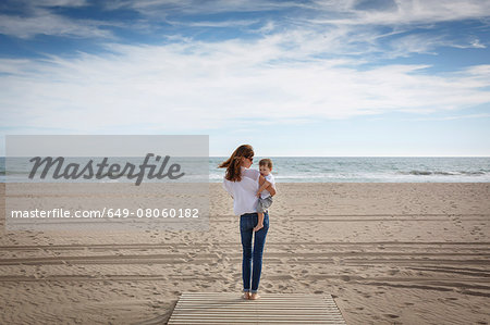 Rear view of mid adult woman carrying toddler daughter on beach, Castelldefels, Catalonia, Spain