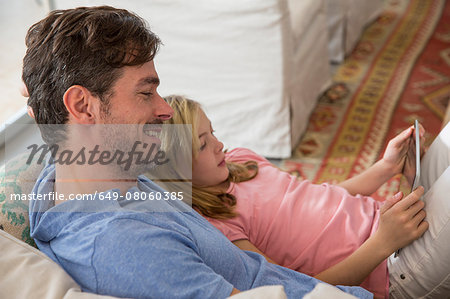 Mature man and daughter reclining on sofa using digital tablet