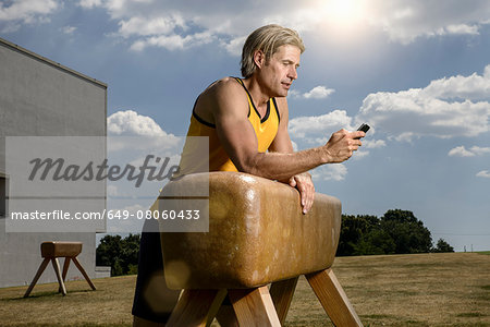 Mid adult male gymnast leaning against vault texting on smartphone