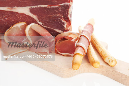 Breadstick wrapped in prosciutto slices on wooden chopping board, big prosciutto piece in background. Culinary prosciutto bright background.